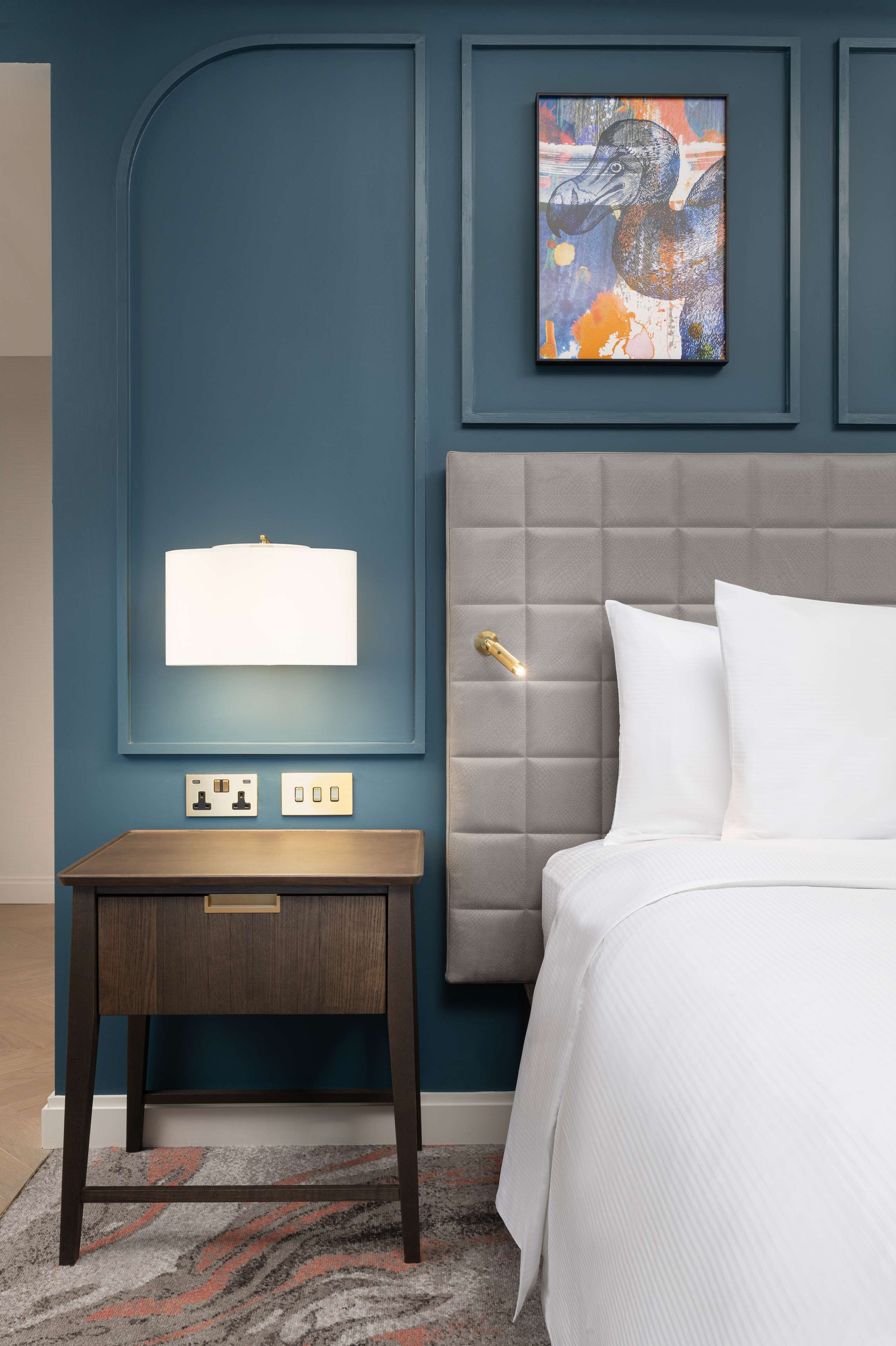 A modern hotel room with a grey tufted headboard, a white bed, a small wooden nightstand with a lamp, and a framed abstract painting on the wall.