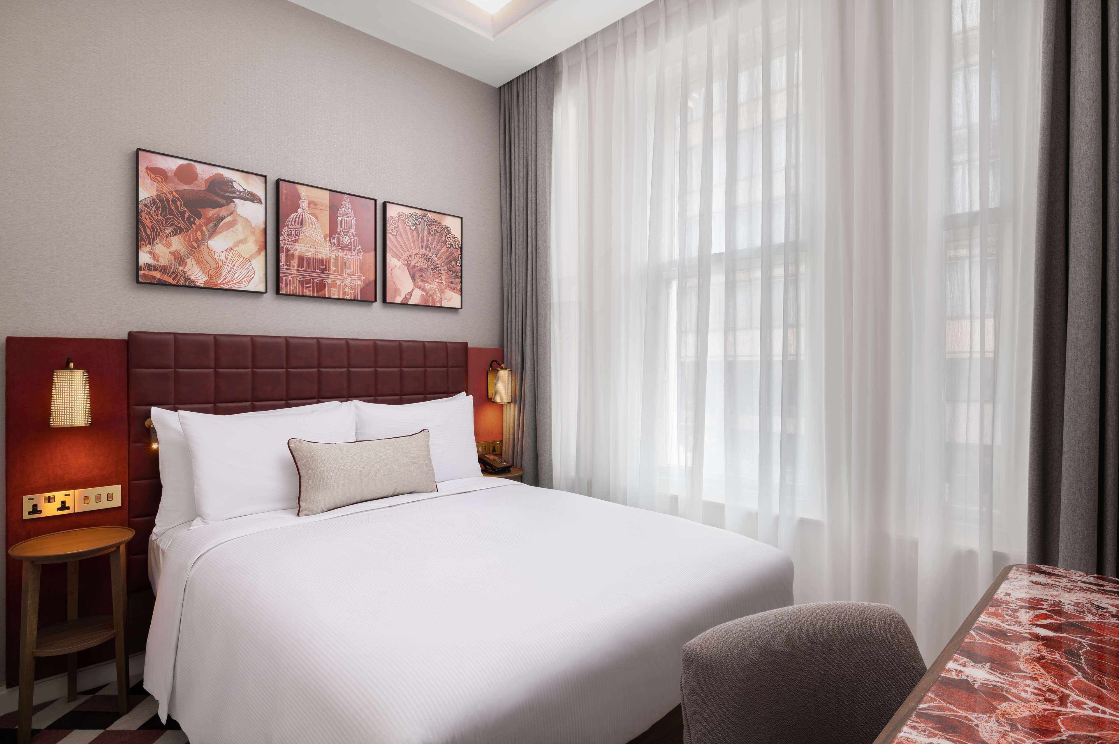 A hotel room with a large bed, two bedside tables, a marble-topped table, and a view of a building through sheer curtains.