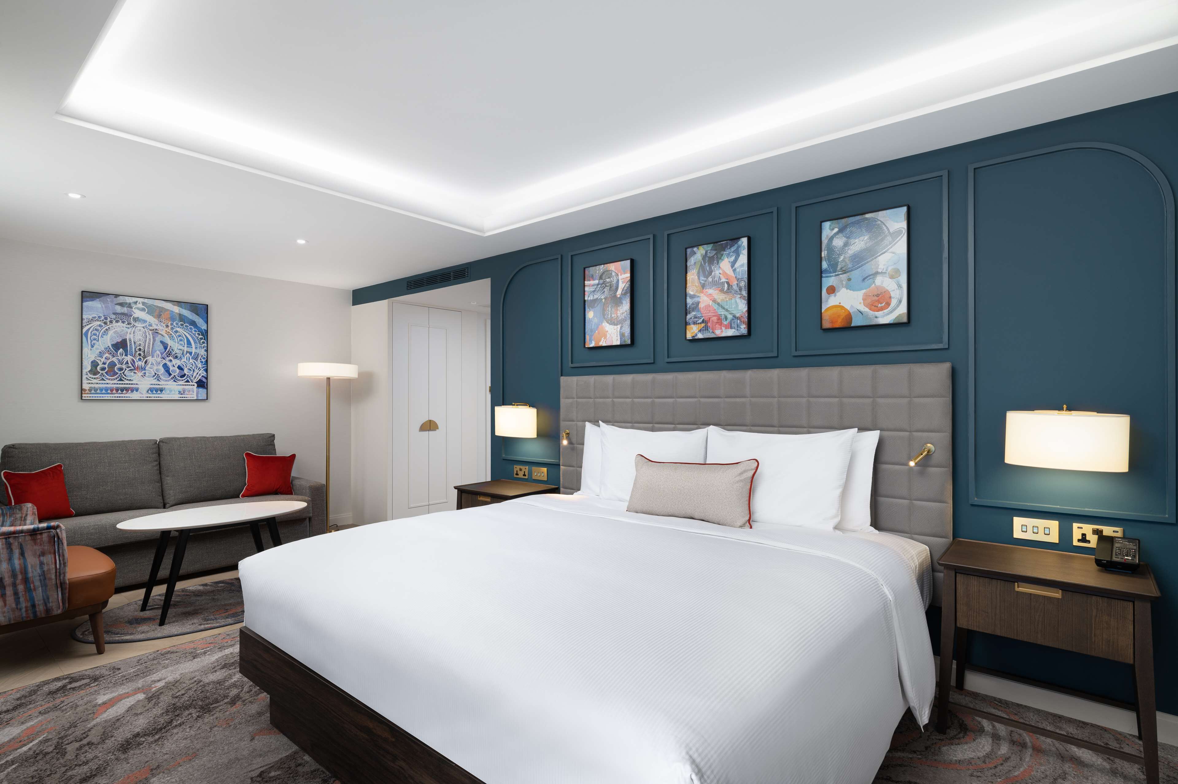 Quarto de hotel moderno com uma cama grande, um sofá, uma pequena mesa e obras de arte nas paredes.