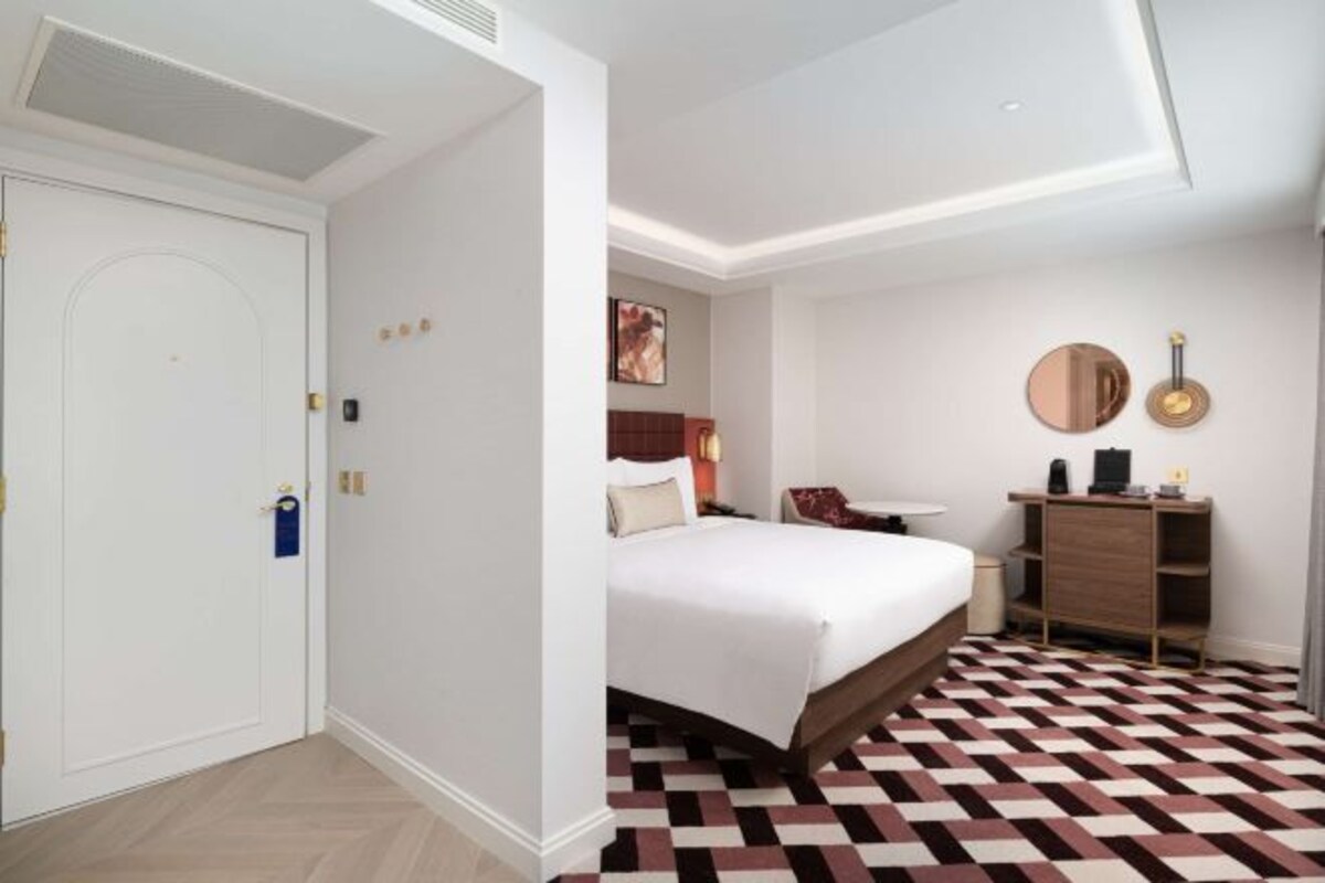 Una habitación de hotel con cama, escritorio, silla, espejo y suelo estampado.