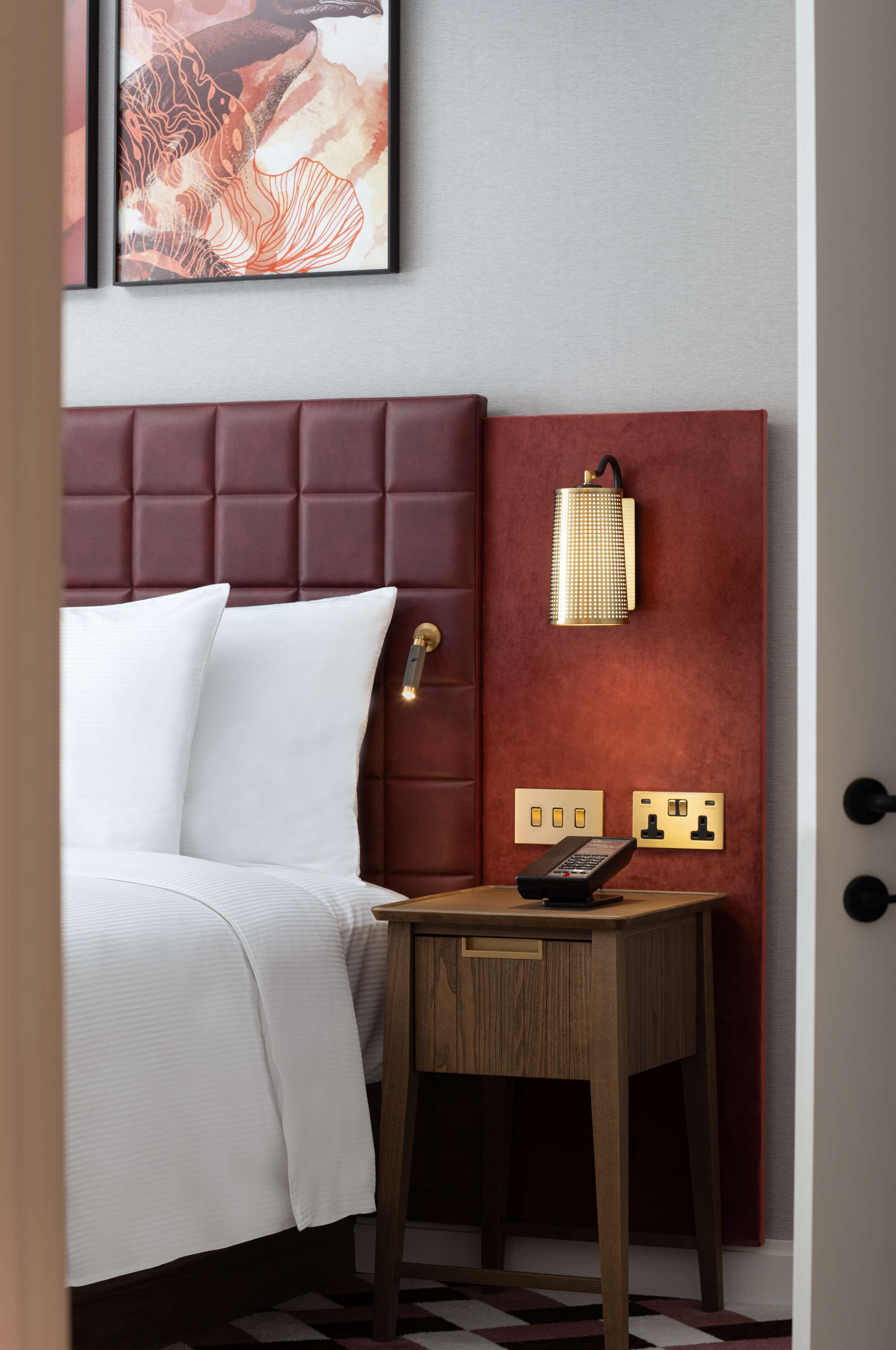 A hotel room with a tufted headboard, a bedside table with a remote control, and a wall-mounted light fixture.