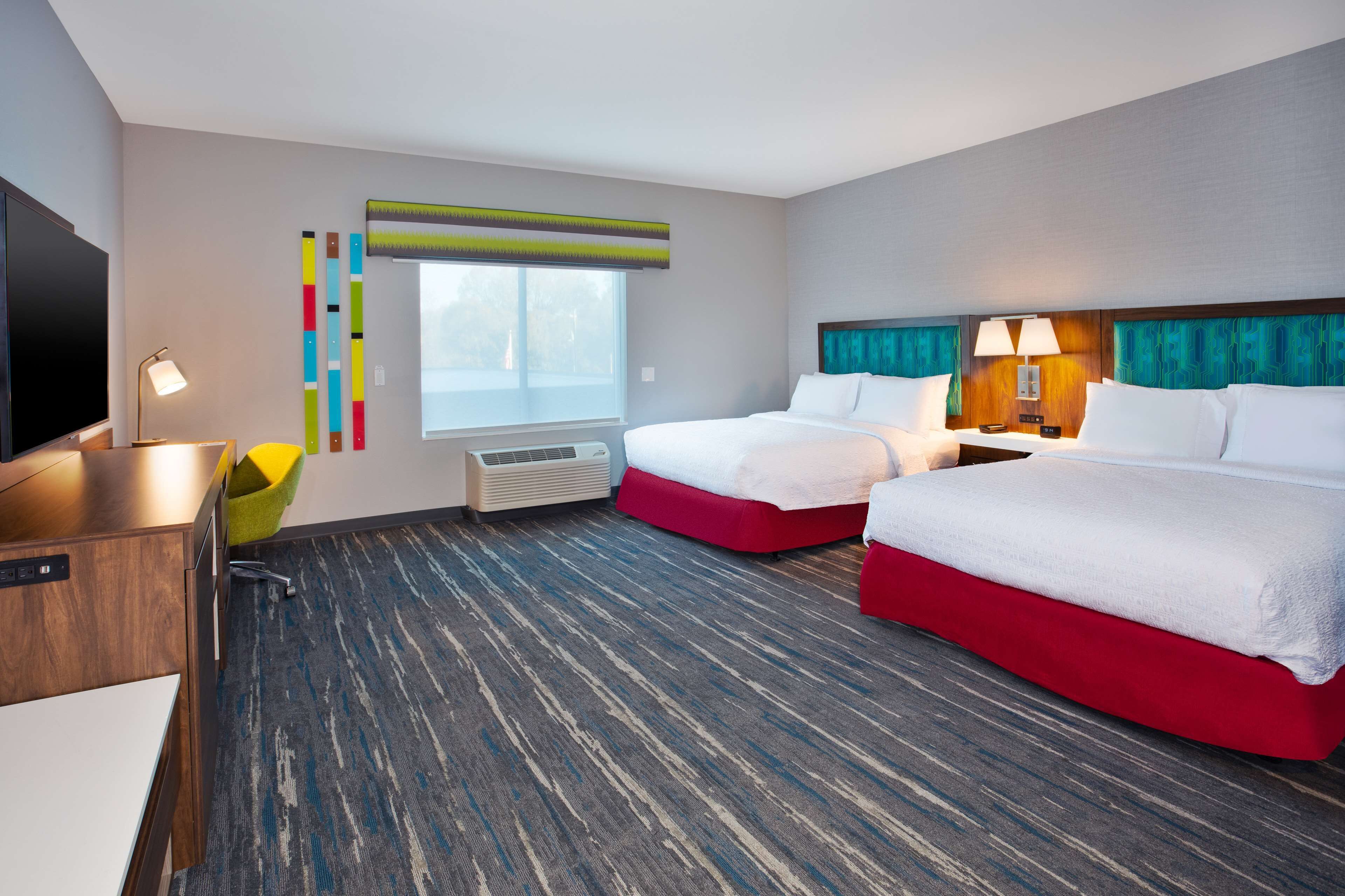 A hotel room with two beds, a desk, a chair, a TV, and a window.