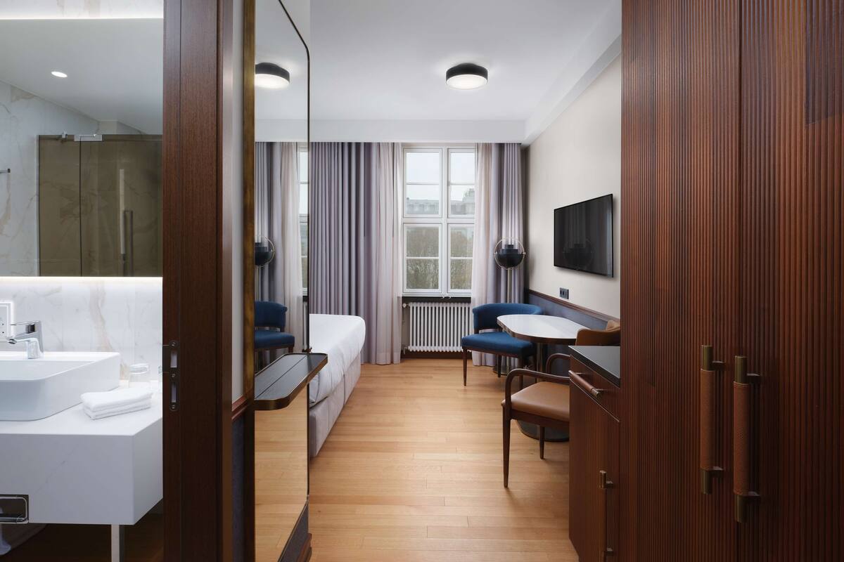 Habitación de hotel con cama, escritorio, silla, televisor, espejo y baño con lavamanos y ducha.
