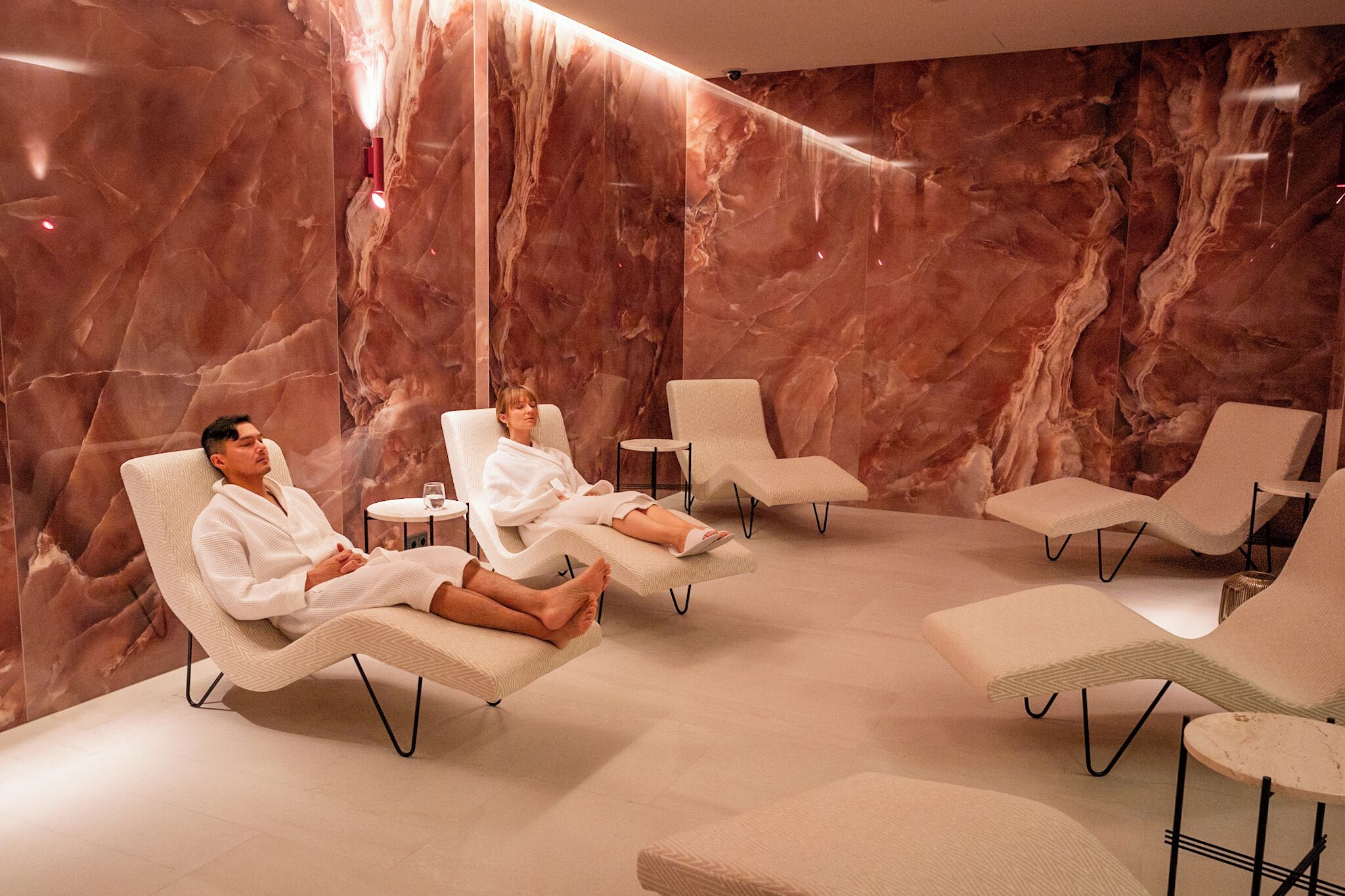 Um espaço de spa com paredes de mármore, cadeiras reclináveis e uma pequena mesa com um copo.