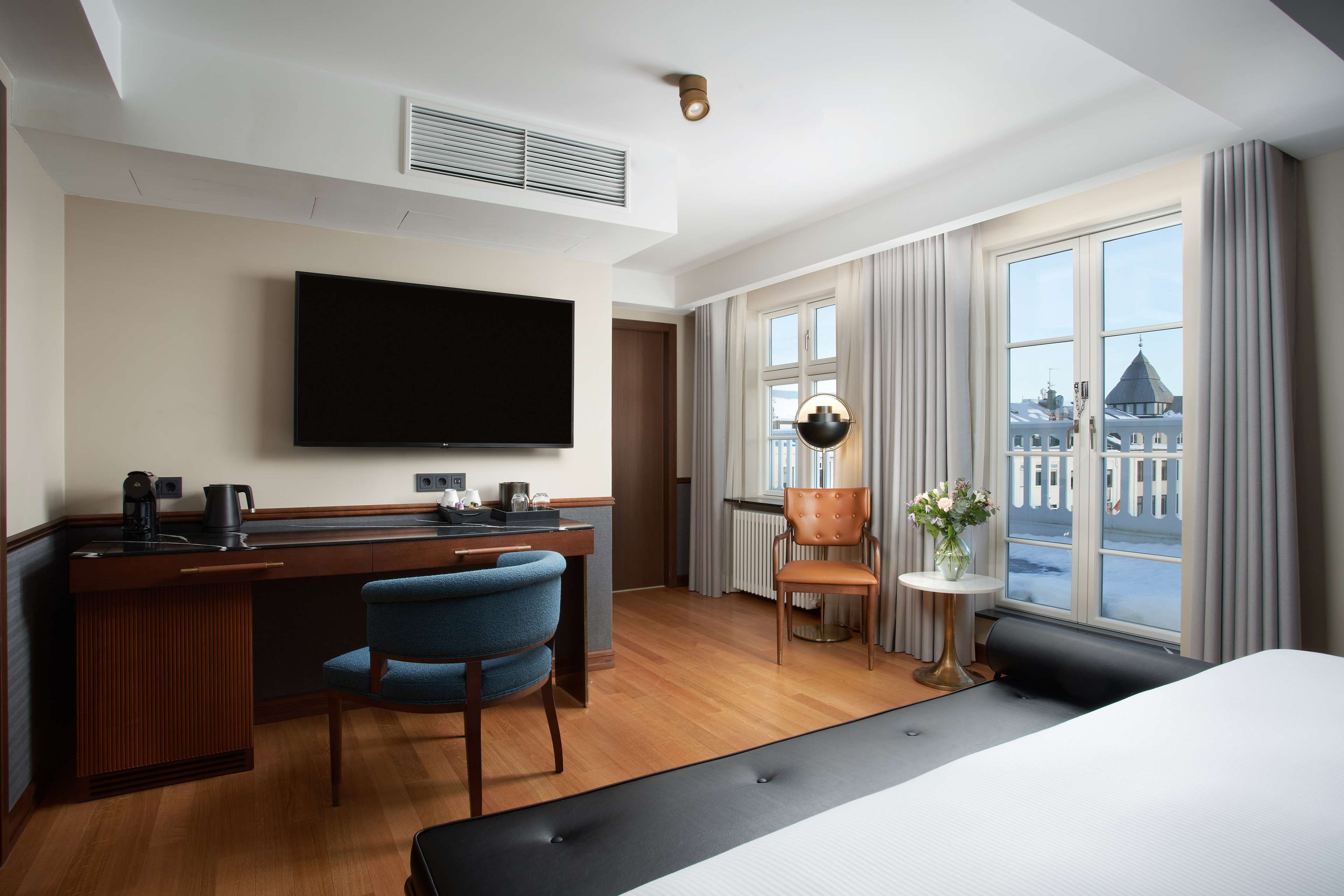 Habitación de hotel con cama, escritorio con televisor de pantalla plana, una silla, una mesita con un florero y ventana con cortinas.