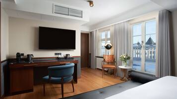 Habitación de hotel con cama, escritorio con televisor de pantalla plana, una silla, una mesita con un florero y ventana con cortinas.