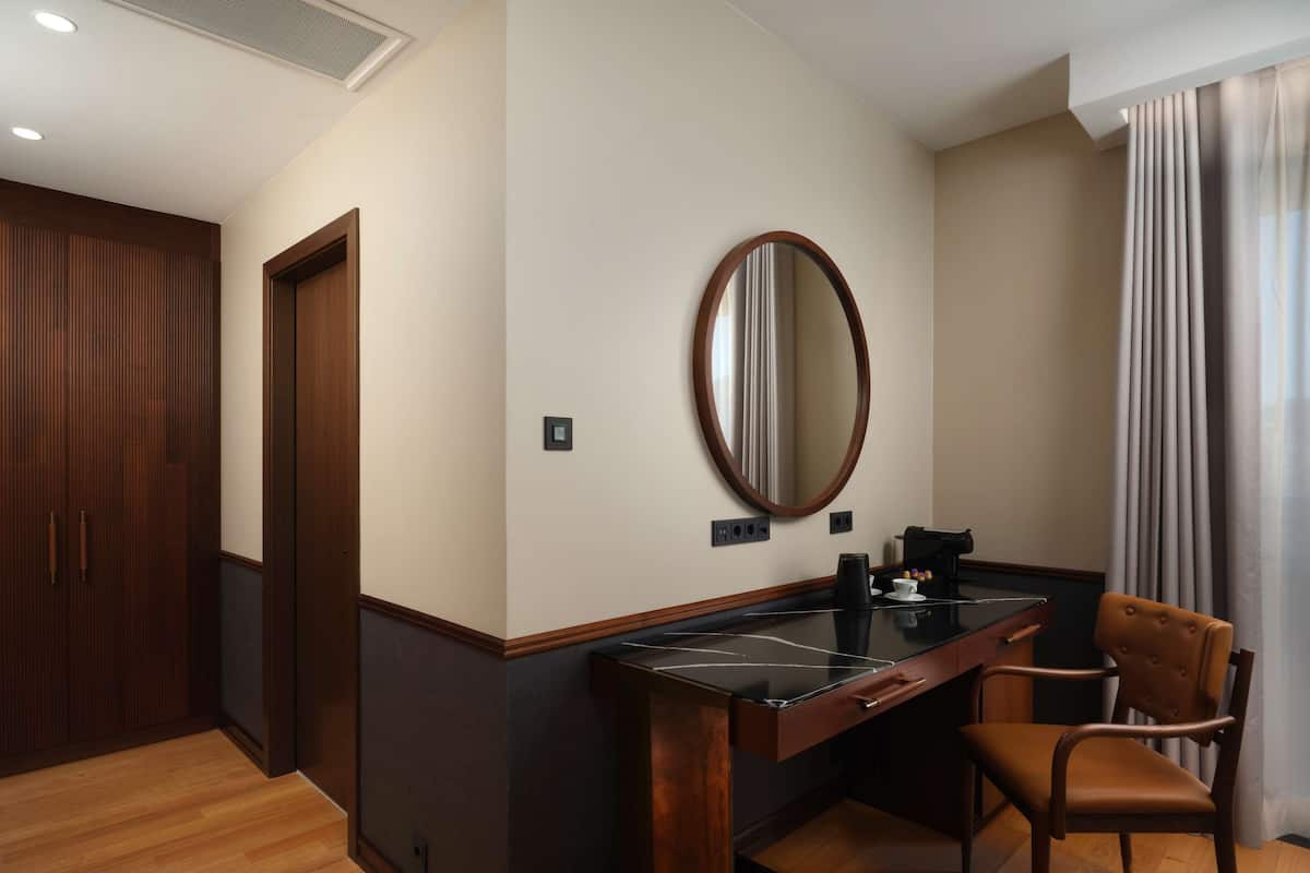 Habitación de hotel con un armario de madera, un escritorio con silla, un espejo redondo y una ventana con cortinas.