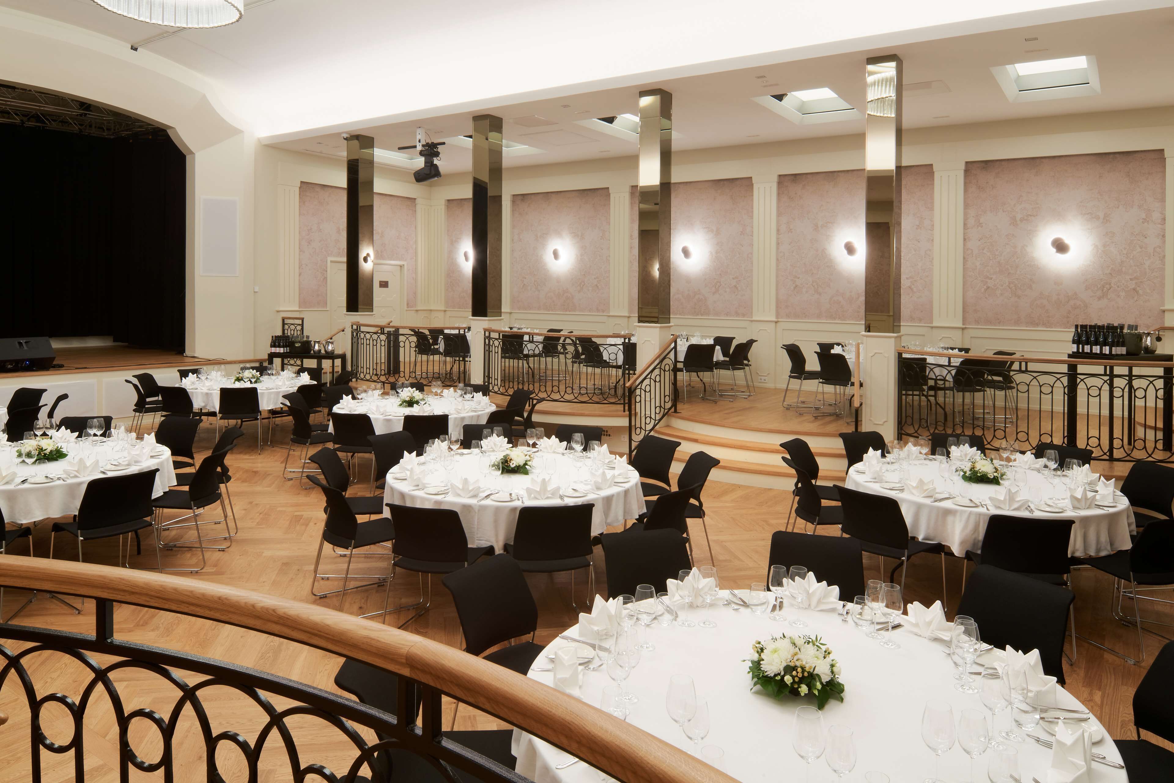 A banquet hall with multiple round tables set for dining, a stage, and a staircase leading to an upper level.