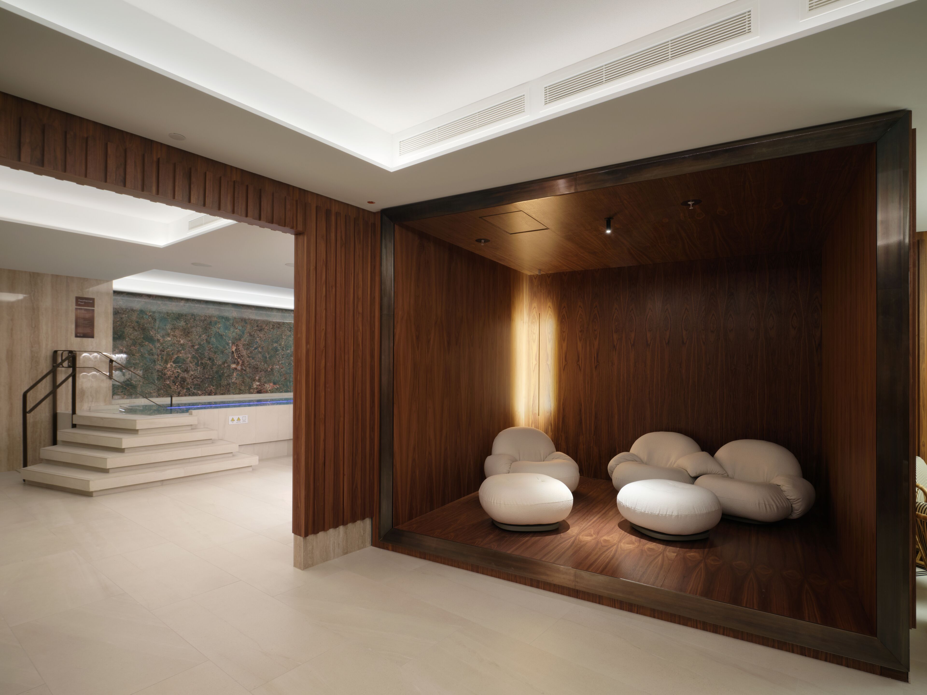A modern spa area with a wooden wall, a seating area with cushioned chairs, and a view of a pool and stairs in the background.