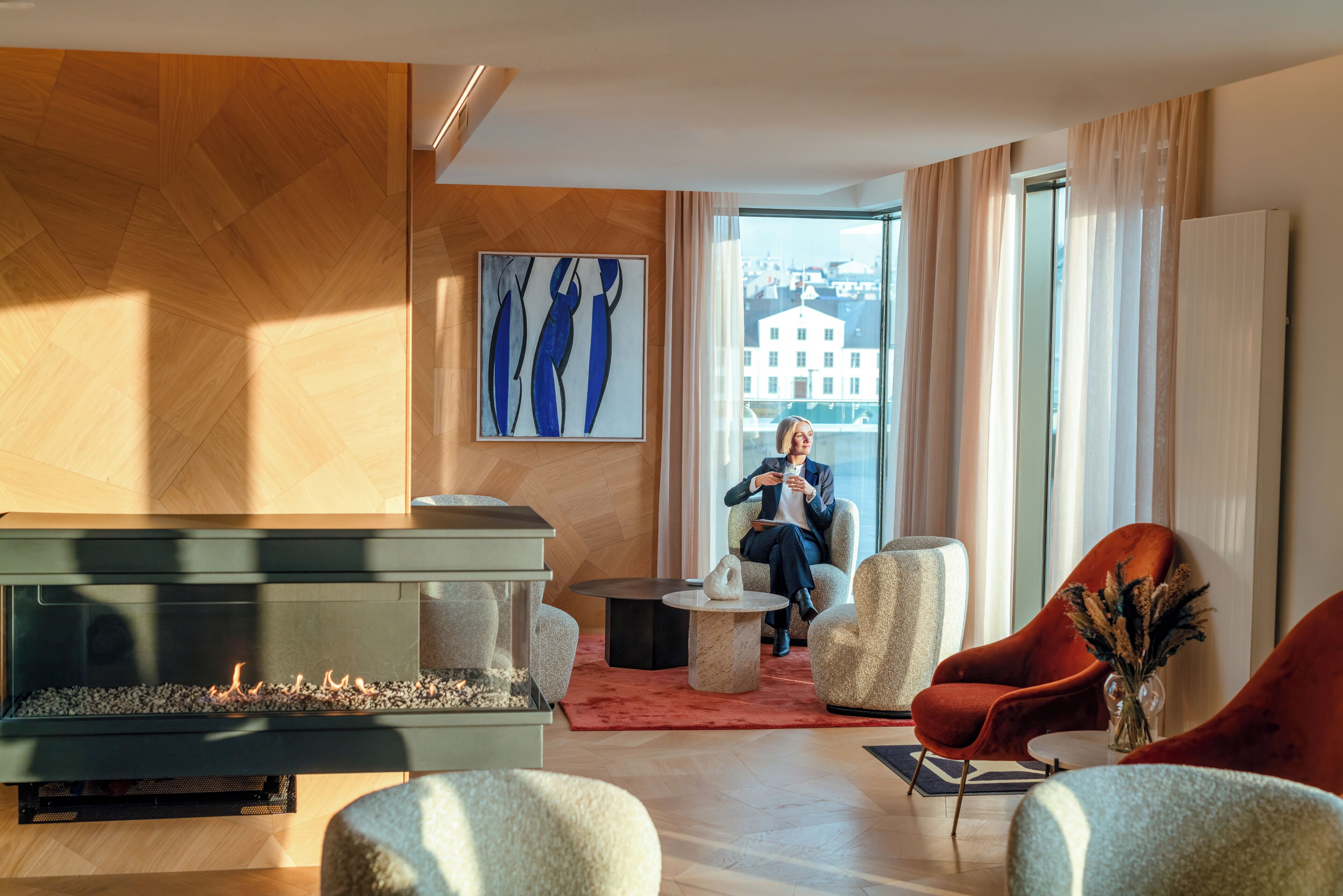A modern living room with a fireplace, comfortable seating, and a large window offering a view of buildings.