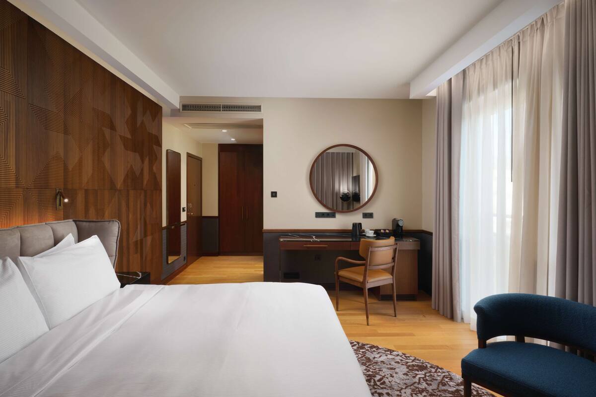 Habitación de hotel con una cama grande, un escritorio con silla, un espejo redondo y vistas al exterior a través de ventanas amplias.