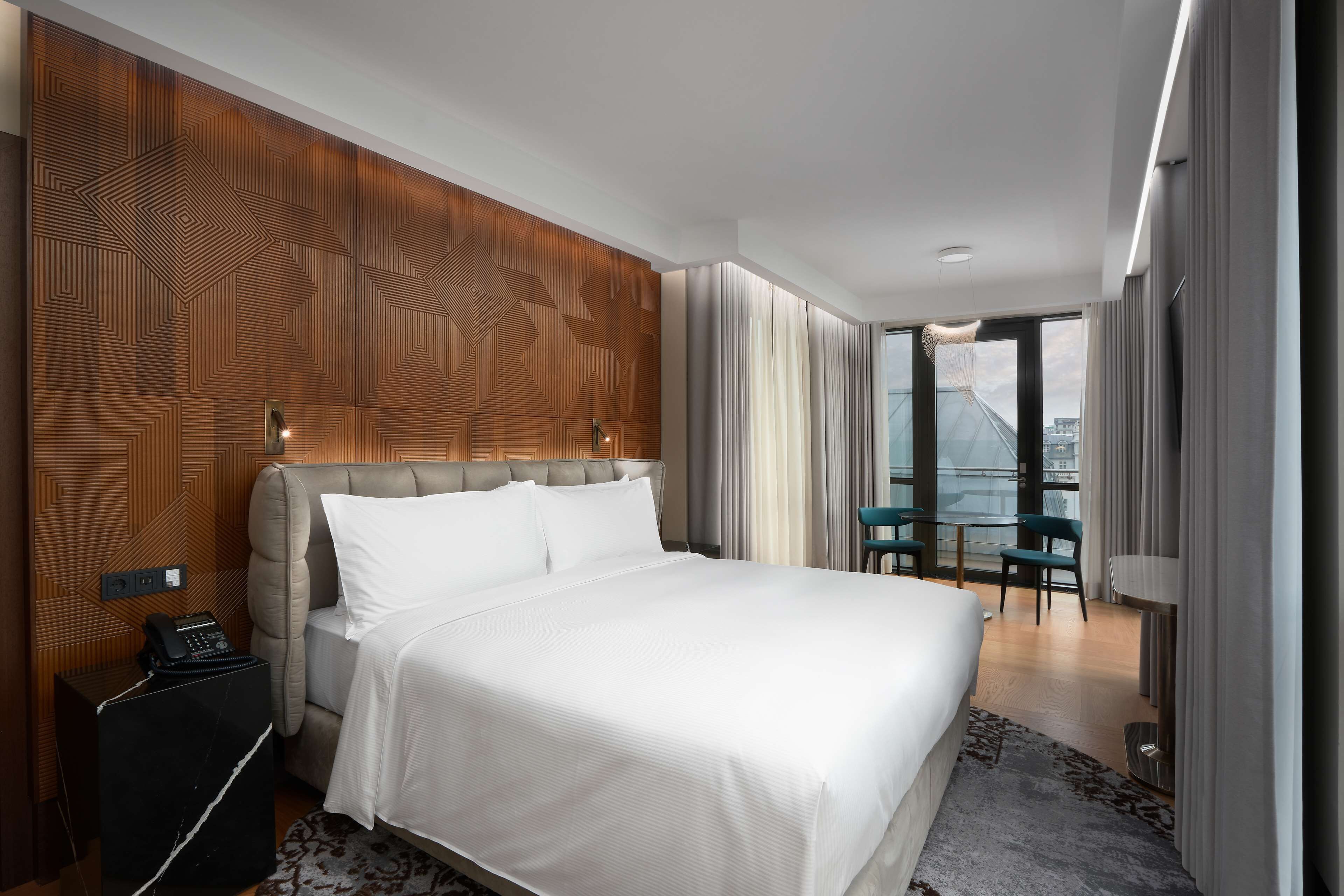 A modern hotel room with a large bed, a wooden accent wall, a small table with chairs, and a view of the city through large windows.