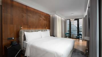 A modern hotel room with a large bed, a wooden accent wall, a small table with chairs, and a view of the city through large windows.