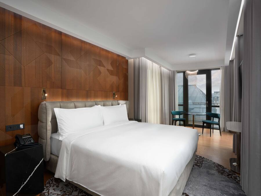 A modern hotel room with a large bed, a wooden accent wall, a small table with chairs, and a view of the city through large windows.