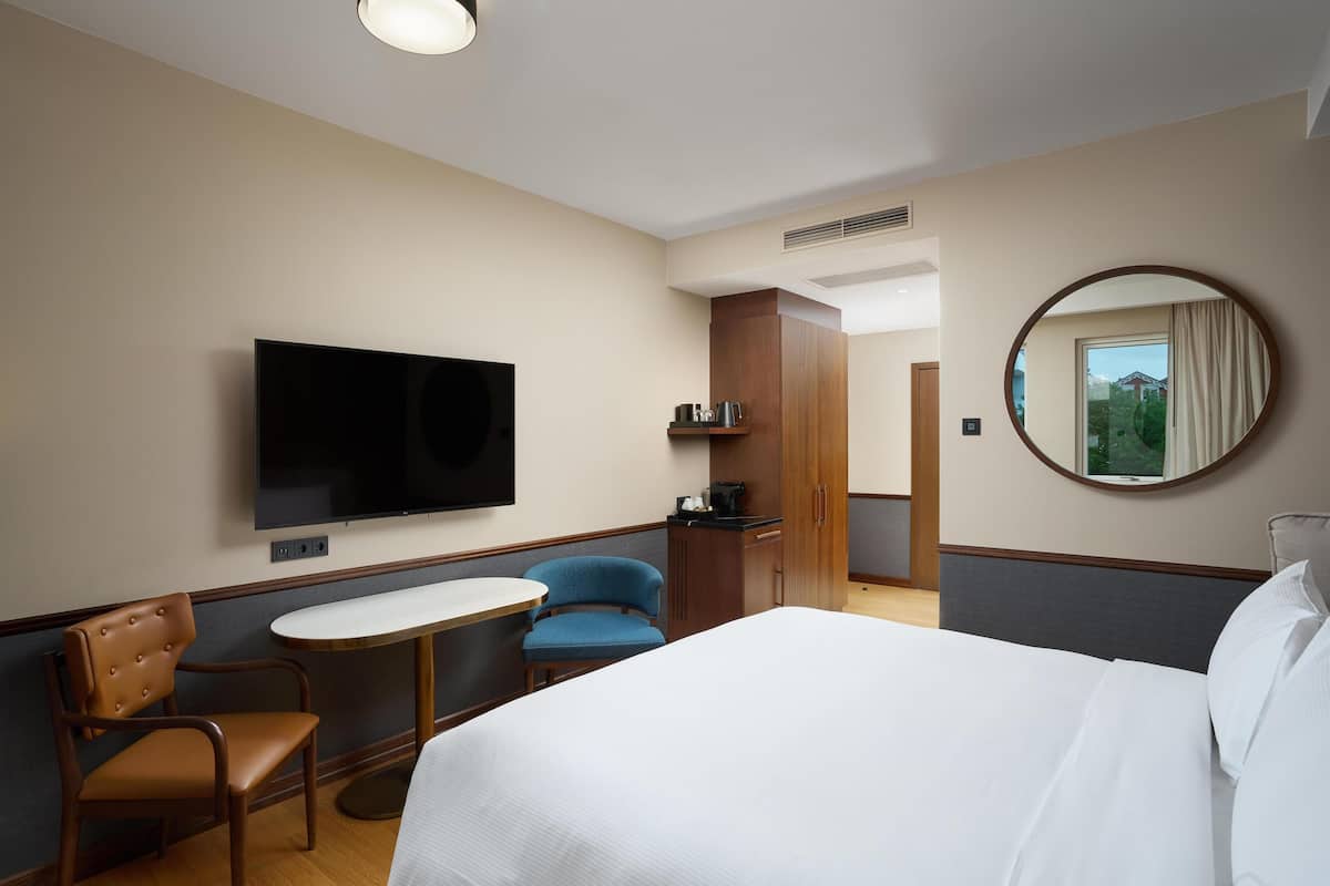 Habitación de hotel con cama, televisión, una mesita, dos sillas, espejo y vista al exterior.