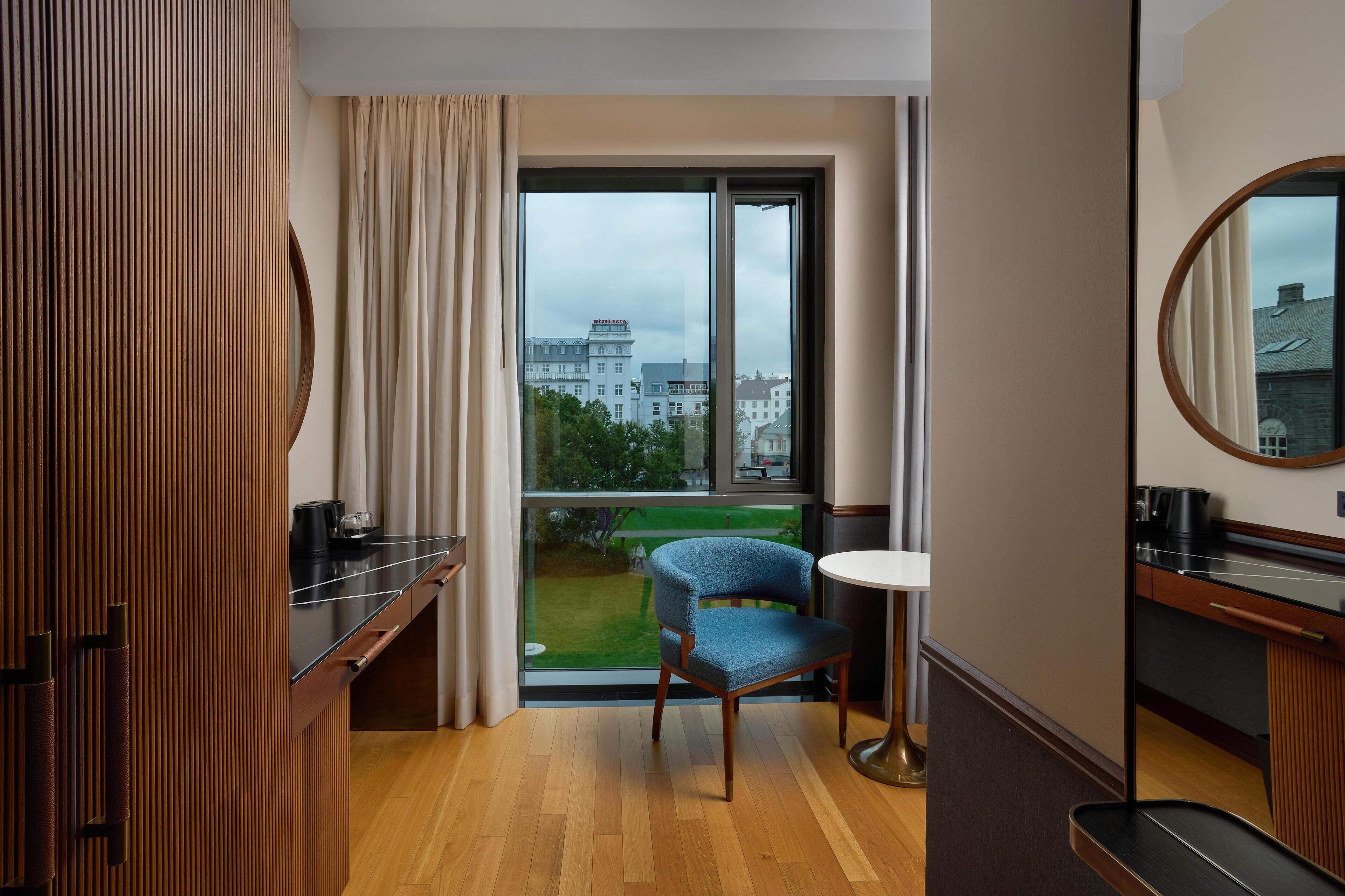 Habitación de hotel con suelo de madera, una silla azul, una pequeña mesa redonda, un espejo y vistas a edificios a través de un ventanal grande.