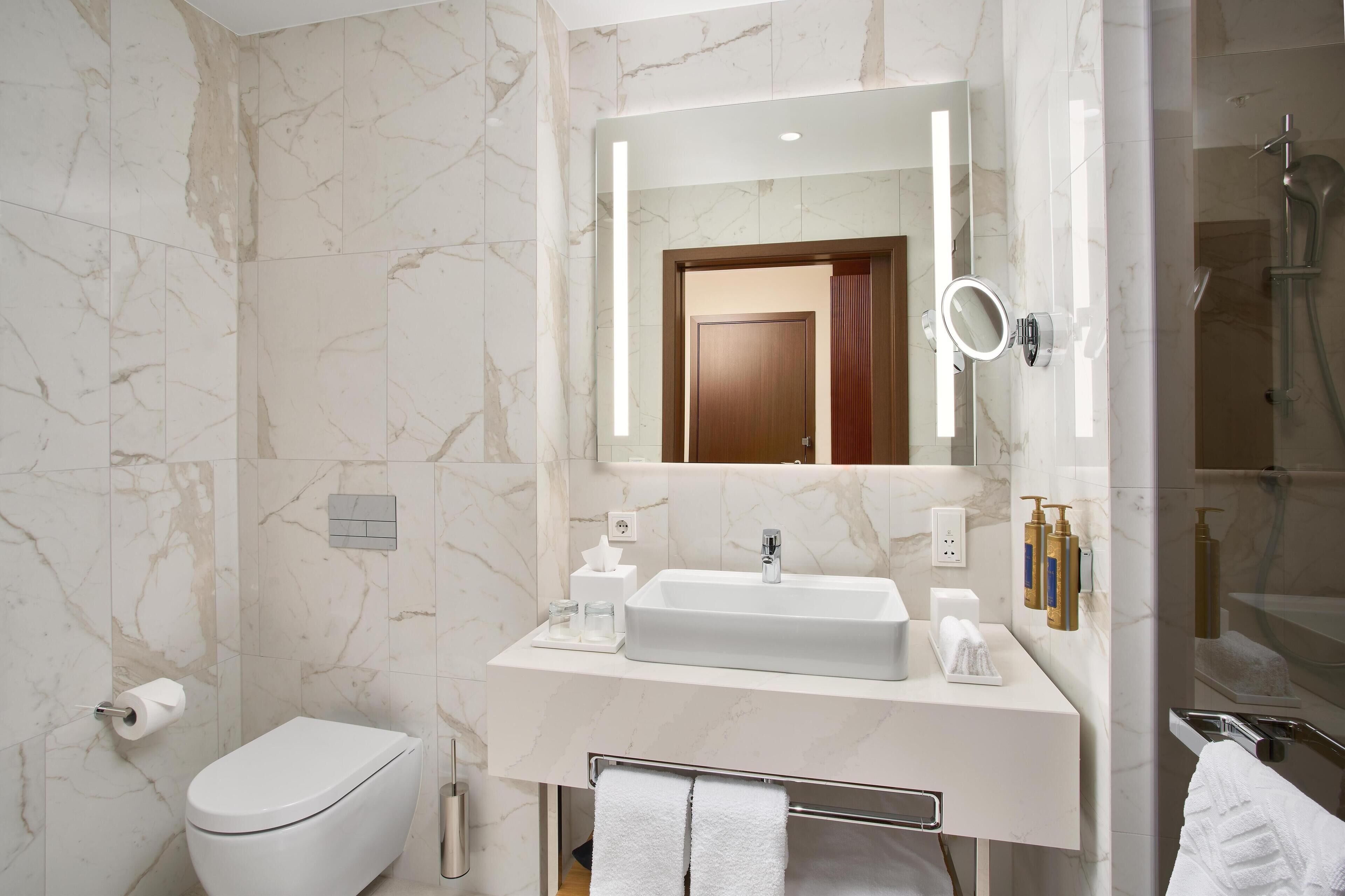 A modern bathroom with a marble wall, a white sink, a toilet, a mirror, and a shower area.