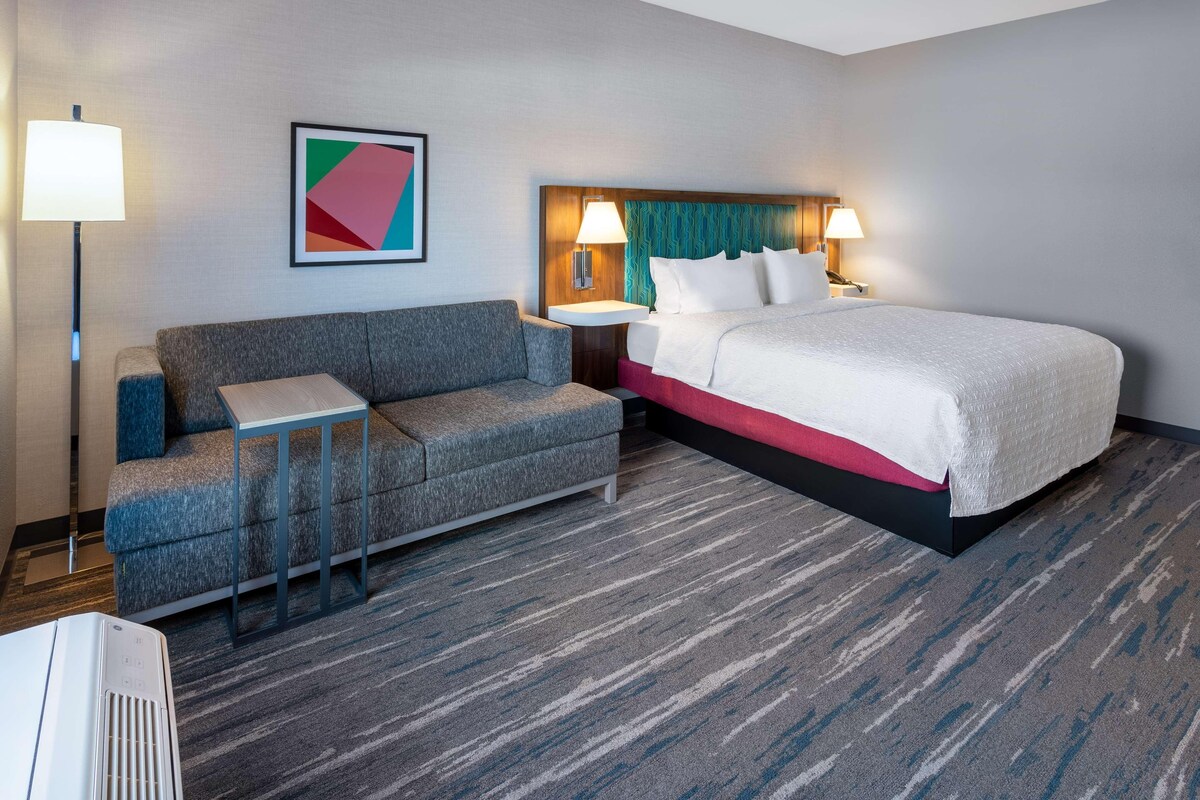 A hotel room with a bed, a sofa, a small table, a lamp, and a wall-mounted artwork.