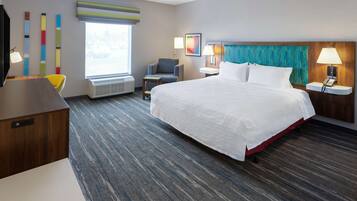 A hotel room with a large bed, a desk, a chair, a small table, a lamp, and a window with blinds.