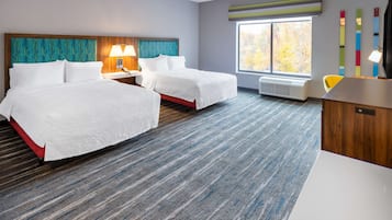 A hotel room with two beds, a desk, a chair, a window with a view, and a wall with colorful decor.