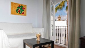 Una sala de estar con un sofá blanco, una mesa de madera oscura con un florero de vidrio, y vista a un edificio desde un balcón con barandilla de metal.