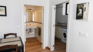 Una habitación con piso de madera, una mesa de comedor con sillas, un área de cocina con lavadora, y un baño con espejo y lavamanos.