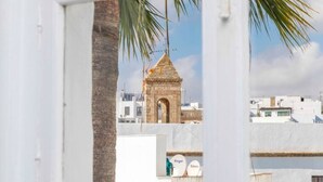 Vista a través de una ventana que muestra una palmera, una pequeña torre de piedra y edificios blancos al fondo.