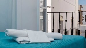 Un letto rifatto con una coperta color petrolio e lenzuola bianche, un balcone con ringhiera nera e una parete bianca sullo sfondo.