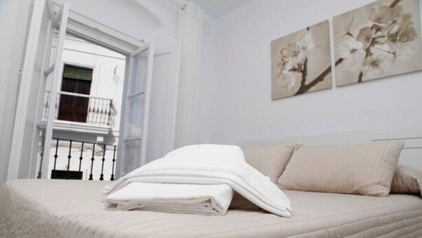 Un letto rifatto con una coperta bianca ripiegata, un cuscino beige e una testiera bianca. Sono presenti due quadri floreali appesi al muro.