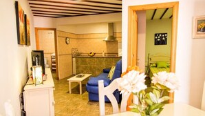 A cozy living room with a blue sofa, a small table, and a vase of flowers. There is a kitchen in the background with a clock and a fruit bowl. - Apartamento El Cotarro B (Vejer de la Frontera)