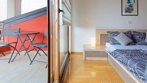 Een slaapkamer met een bed, nachtkastje en een bijzettafeltje. Er is een raam met uitzicht op een buitenruimte met een tafel en stoelen.