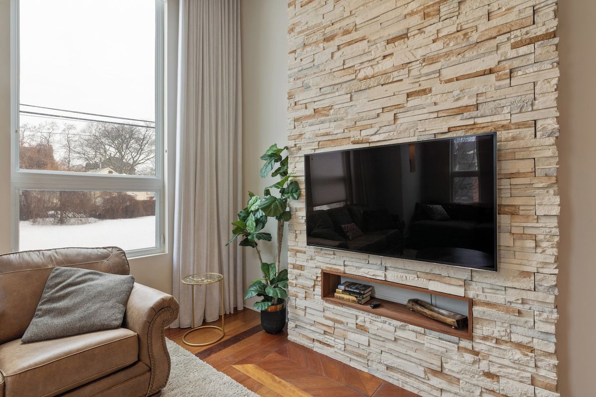 Ein Wohnzimmer mit Steinwand, Flachbildfernseher, Ledersofa, Topfpflanze und einem Fenster mit Vorhängen.