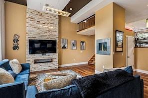 A living room with a stone fireplace, a flat-screen TV, and a staircase leading to an upper level.