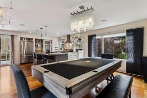 A modern kitchen with a pool table, dining area, and large windows.
