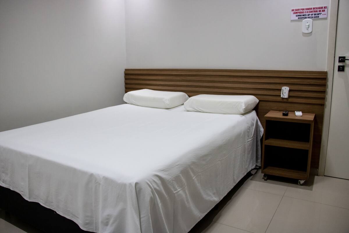 Una cama bien tendida con sábanas blancas, un cabecero de madera y una mesita de noche con un control remoto.