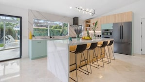 A modern kitchen with a central island, bar stools, a stainless steel refrigerator, and a large window overlooking a garden. - Colorful Modern Luxury w/ Stunning Mountain Views (Palm Springs)
