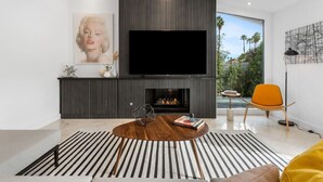 A modern living room with a flat-screen TV, a fireplace, and a large window offering a view of palm trees. - Colorful Modern Luxury w/ Stunning Mountain Views (Palm Springs)