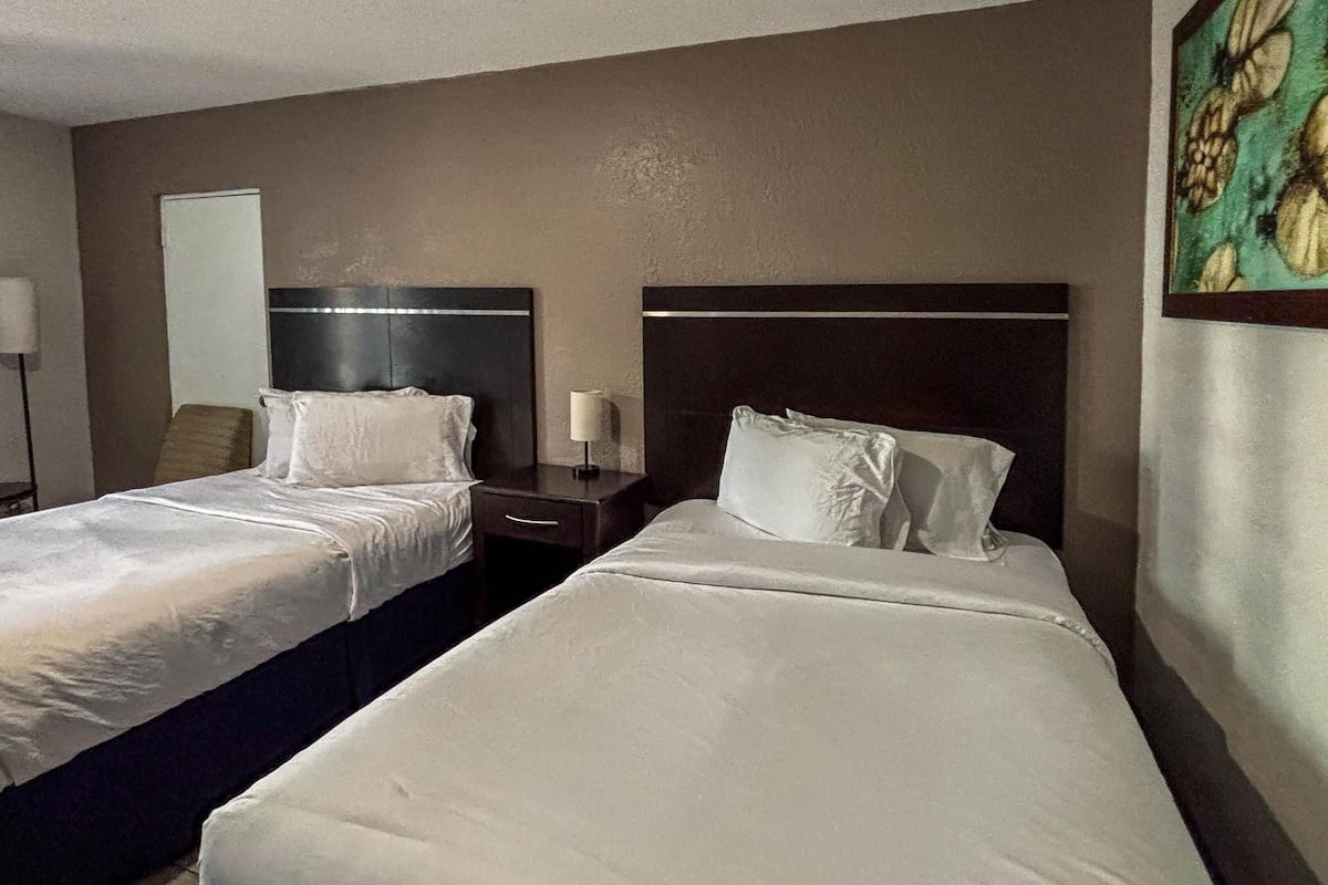 A hotel room with two single beds, a nightstand with a lamp, a wall-mounted mirror, and a painting on the wall.