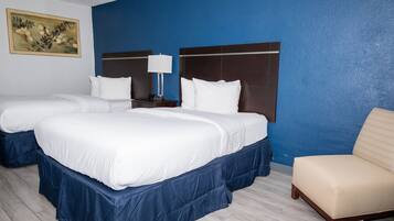 Quarto de hotel com duas camas, uma parede de destaque azul, uma poltrona bege e uma mesa de cabeceira com luminária.