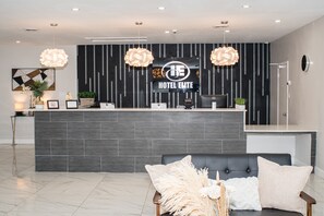Un comptoir d’accueil d’hôtel au design moderne, comprenant un mur à rayures noires et blanches, un logo au-dessus du comptoir et un espace d’accueil avec un canapé et des coussins décoratifs.