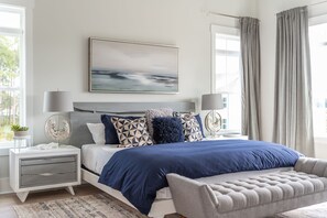 Quarto moderno com cama grande, mesas de cabeceira, um banquinho, um quadro e vista para o exterior.