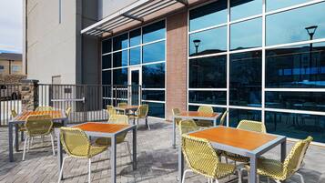 An outdoor patio area with tables and chairs, large windows, and a brick wall.