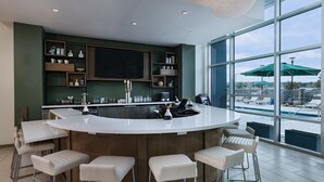A modern kitchen with a large island, bar stools, and a view of a swimming pool through large windows. - Element by Marriott Dallas Las Colinas (Irving)