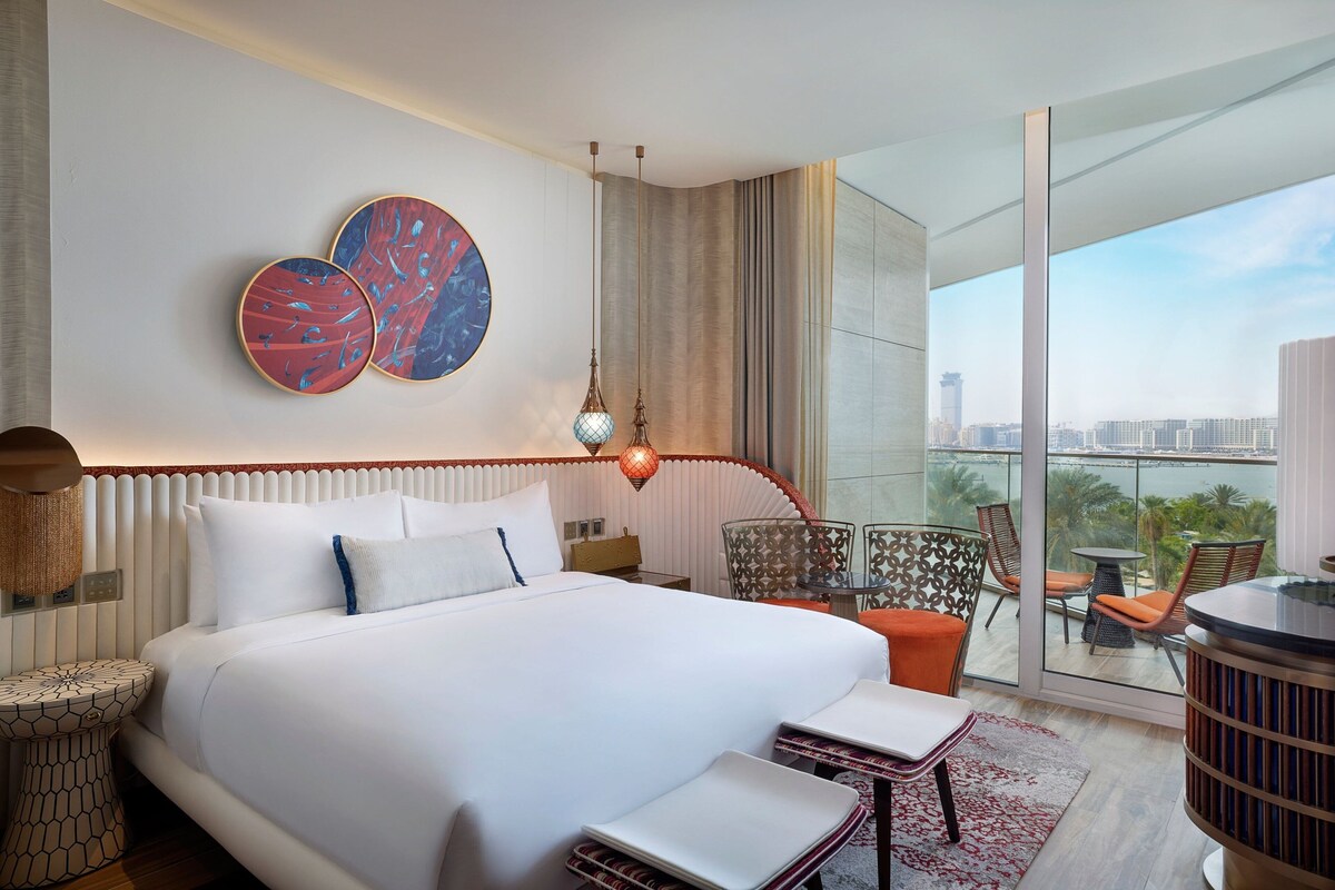 A modern hotel room with a large bed, a balcony with a city view, and two abstract wall art pieces.