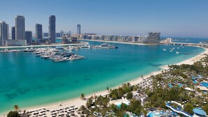 A coastal cityscape with a marina, high-rise buildings, and a beach area with umbrellas and a pool.