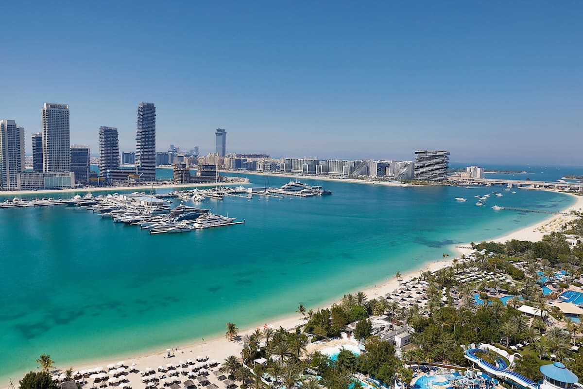 Un paysage urbain côtier avec un port de plaisance, des immeubles de grande hauteur et une plage agrémentée de parasols et d’une piscine.