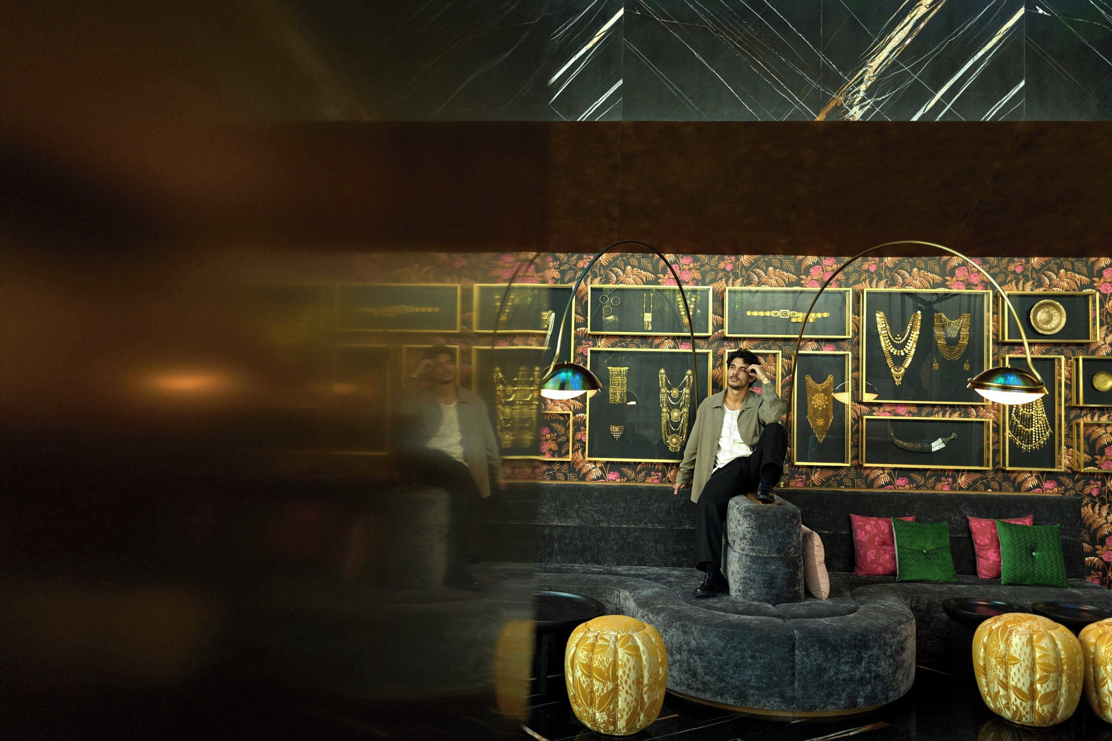 A man sitting on a grey sofa in a room with a wall decorated with framed jewelry. There are two hanging lamps and a patterned carpet.