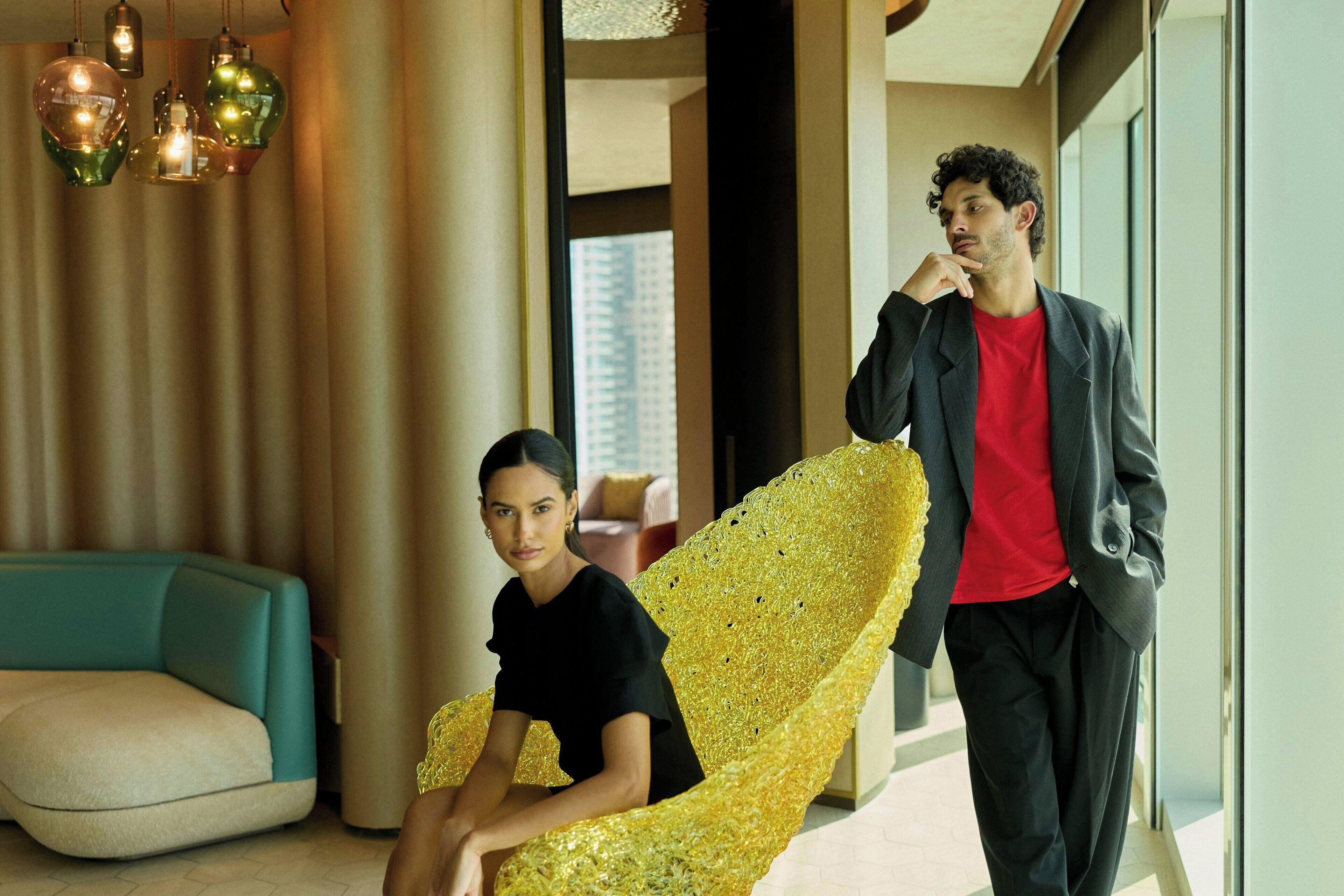 Ein modernes Interieur mit einem türkisfarbenen Sofa, einem goldenen Sessel und einer Person in rotem Hemd und schwarzer Jacke.