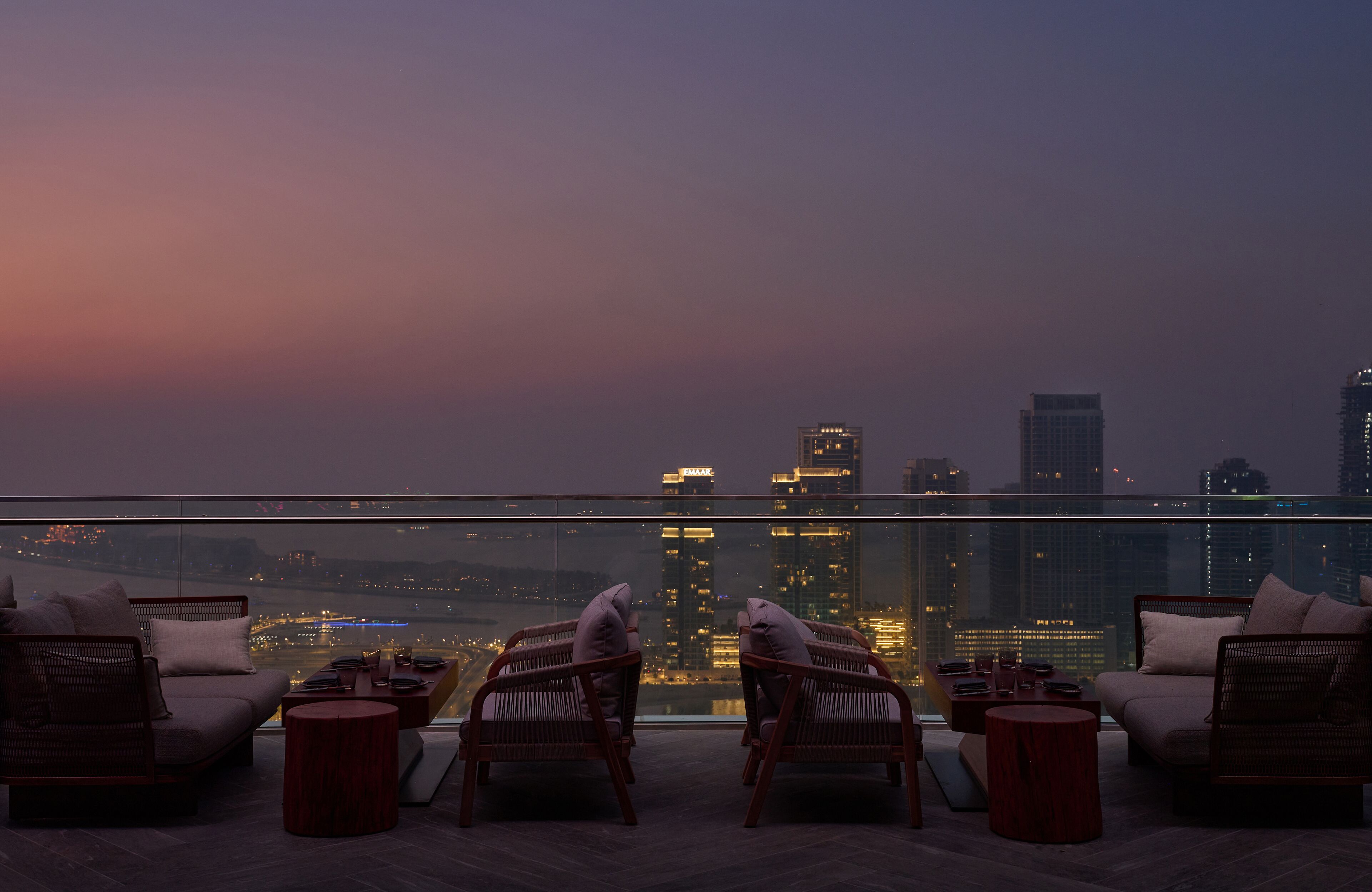 A rooftop lounge with wicker furniture and a city view at dusk.