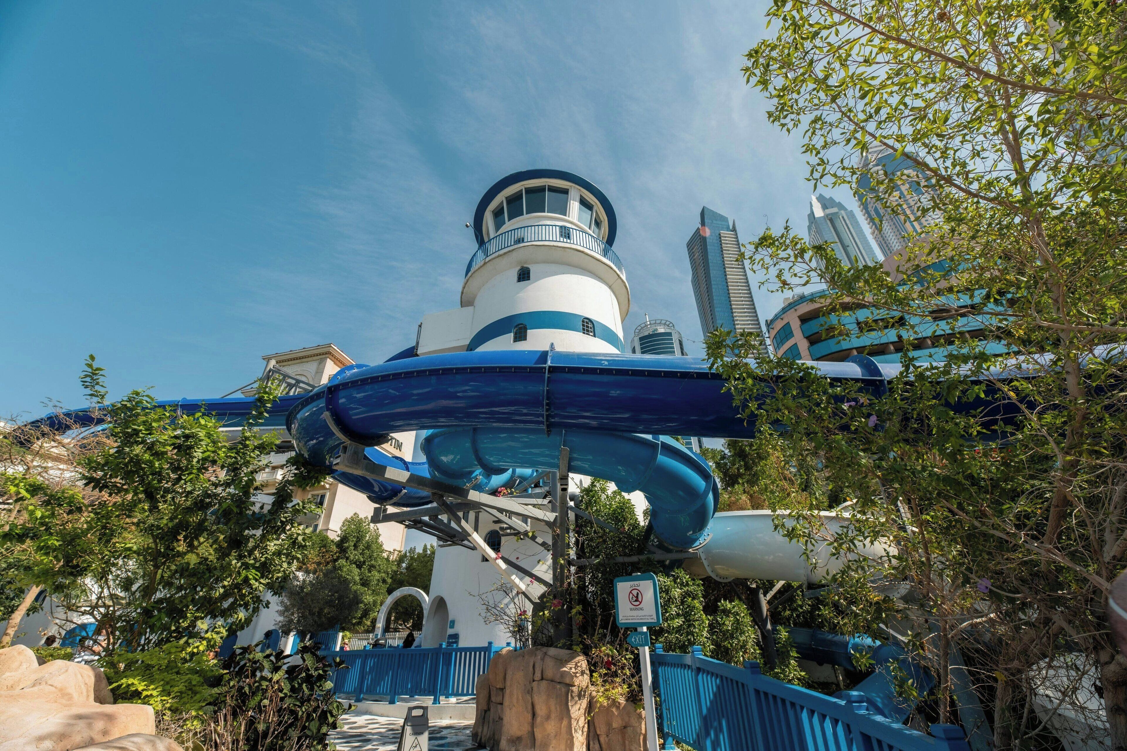 A water park with a large water slide, a lighthouse structure, and modern buildings in the background.